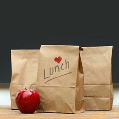 Three brown lunch bags sit next to a red apple. One brown bag has “Lunch” written on it with a small heart.