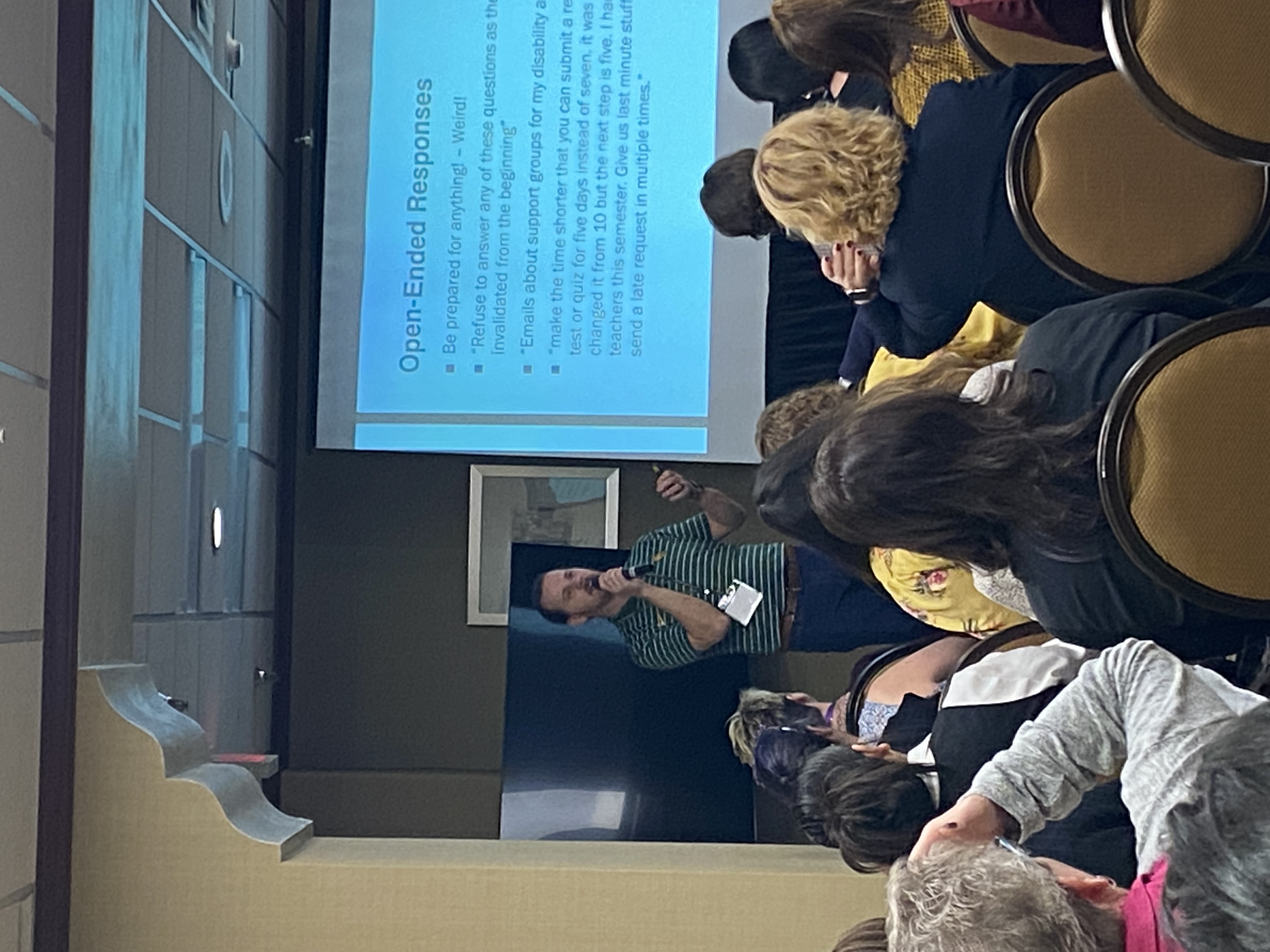 Conference photograph of session with seated attendees and presenter in front of large display board with PowerPoint displayed