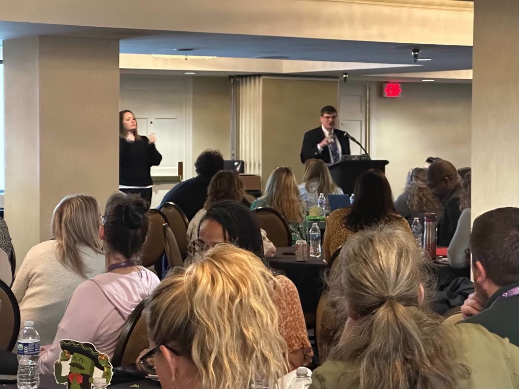 Room full of people for the Keynote address. Man in suit presenting to audience with an ASL interpreter to the left side of the podium